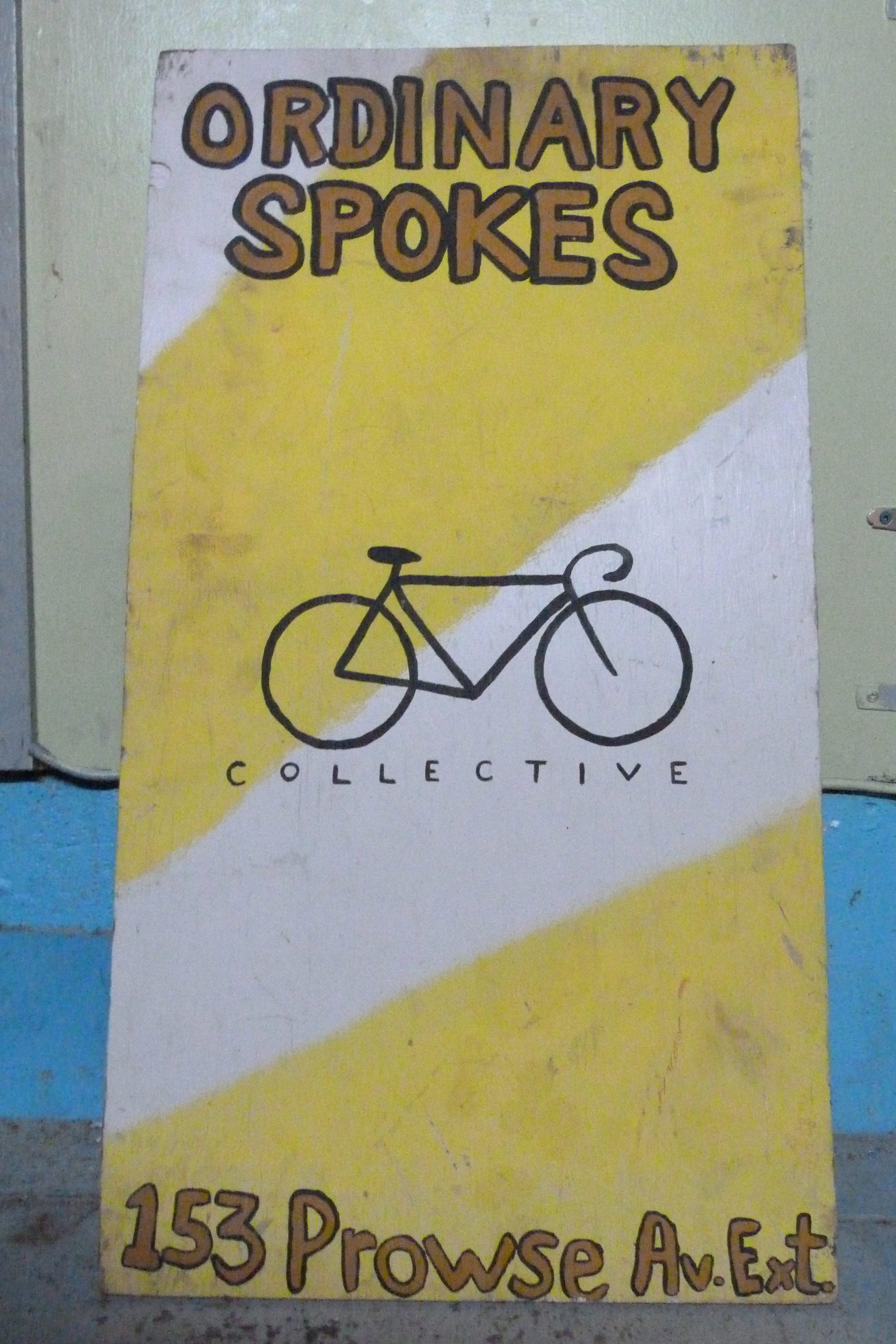 Yellow signboard. Text reads Ordinary Spokes. Picture of a bicycle with the word "Collective" underneath.
